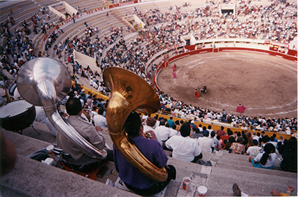 Two men with tubas sit in the stands of a bullfighting arena with other spectators