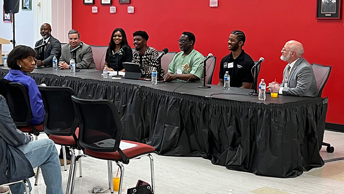 A panel of seven people seated at a long table with microphones during the opening of the Africana Studies Collection, addressing an audience in a room with red walls and framed plaques.