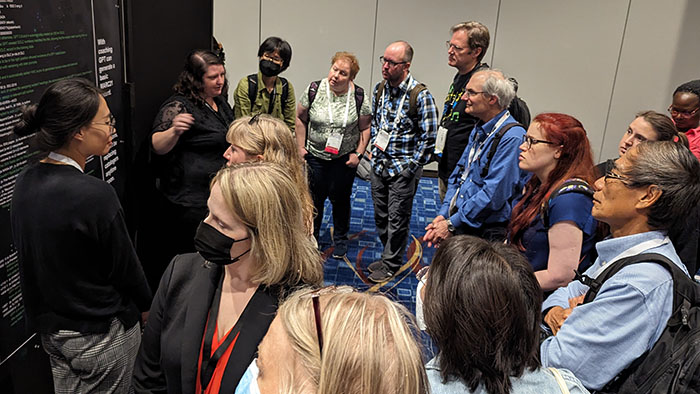 A group of people gathered around a SDSU librarian at a conference, listening attentively to a discussion near a large display board with text.