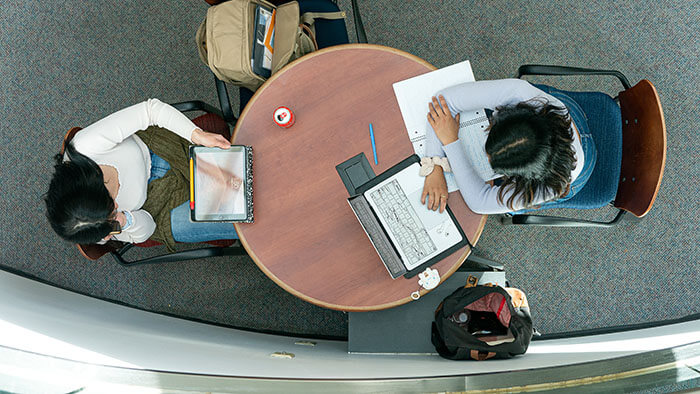 Overhead view of two students working at a table, one on a laptop, the other on a tablet