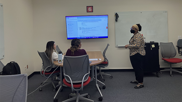 A librarian instructs two students in a classroom with a large screen displaying a webpage, a table, chairs, and a whiteboard in the background.