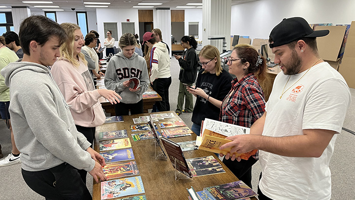 A group of SDSU students browse through comic books displayed on tables in Special Collections. Some students are engaged in conversation, while others are reading or using their phones. The comics are arranged neatly, with colorful covers facing up.