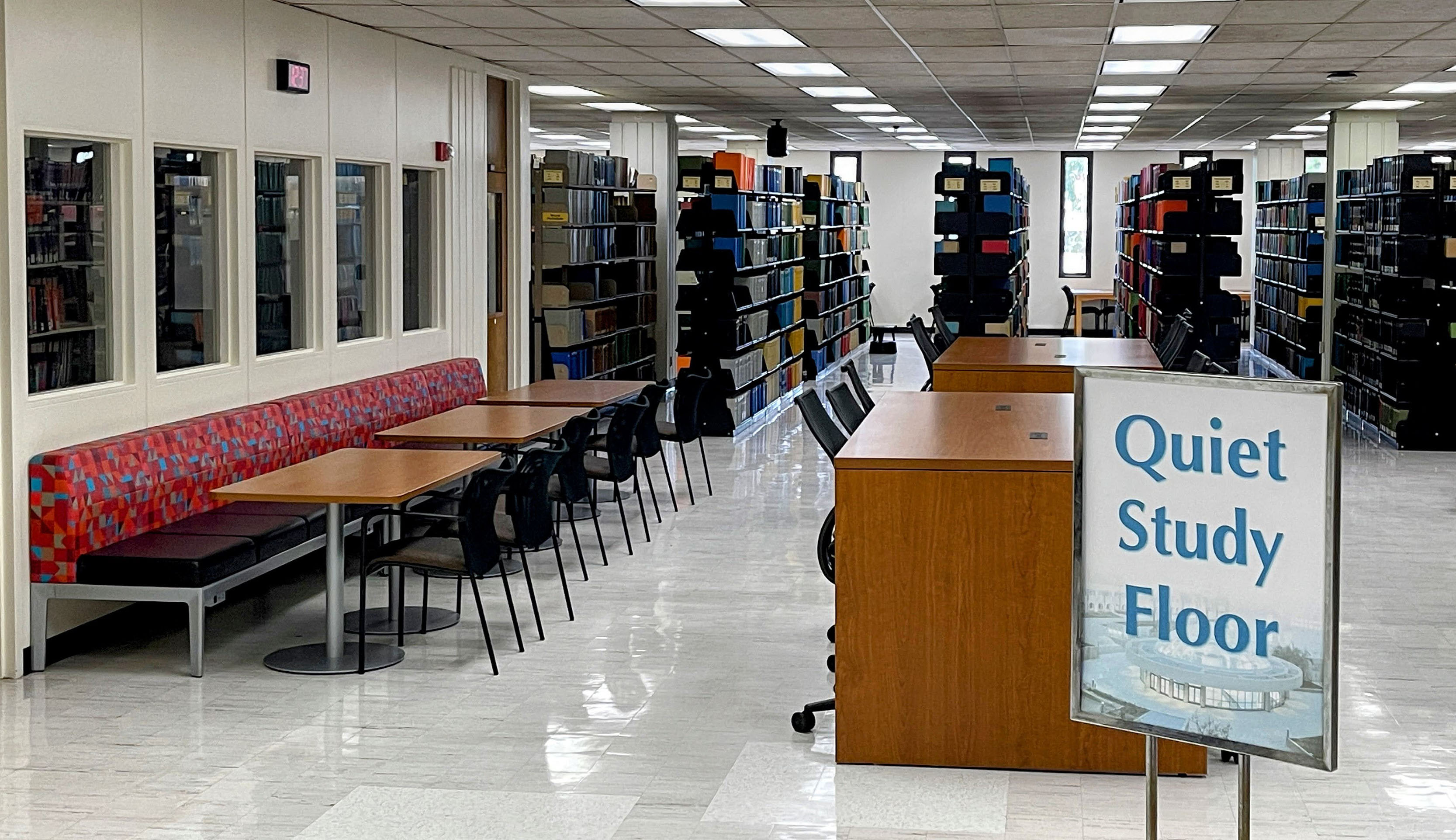  An image of a quiet study space in a library with empty tables and chairs, a sign that says Quiet Study Floor, and bookshelves in the background.