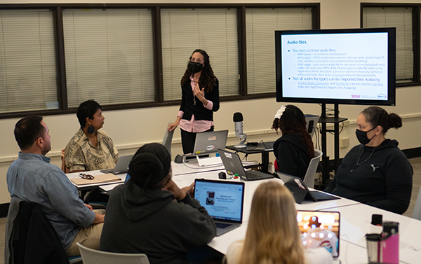 Adults seated around a small table with professor standing next to a slideshow that reads Audio Files